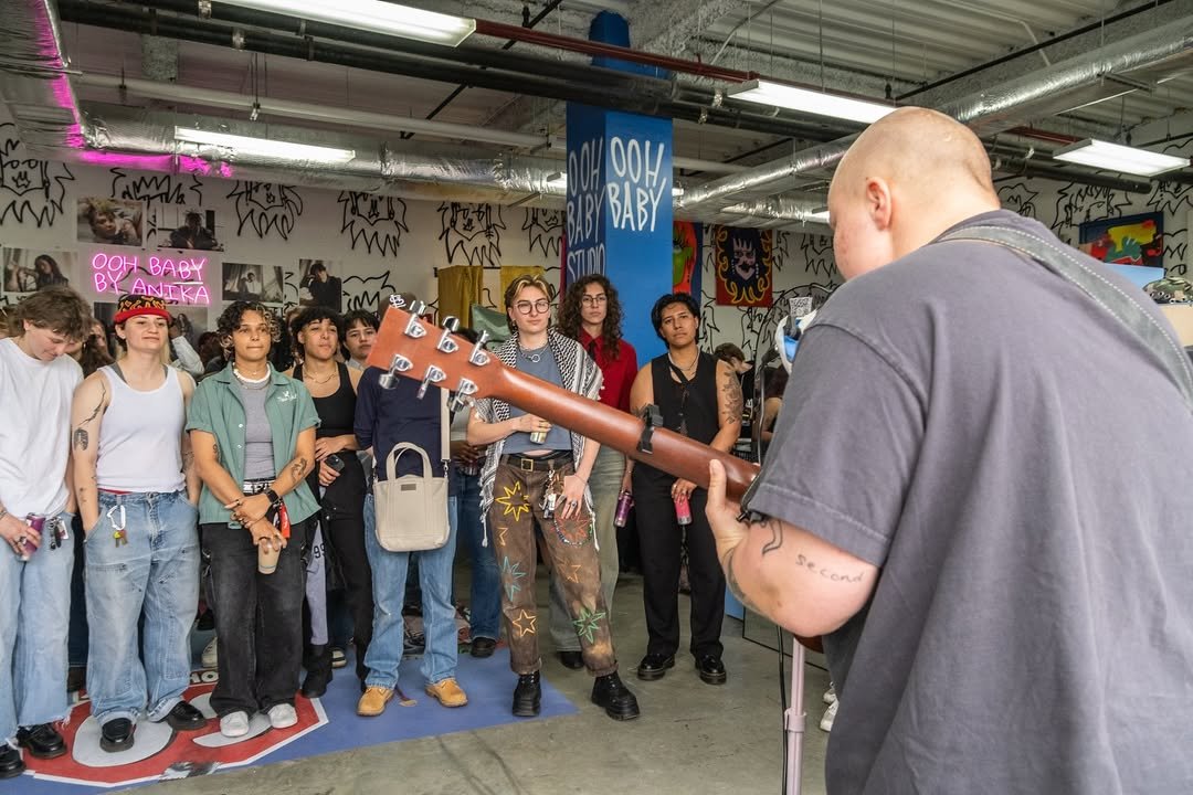 A person with a bald head playing an acoustic guitar in front of a group of young people in a music or art venue, with graffiti and artwork on the walls.